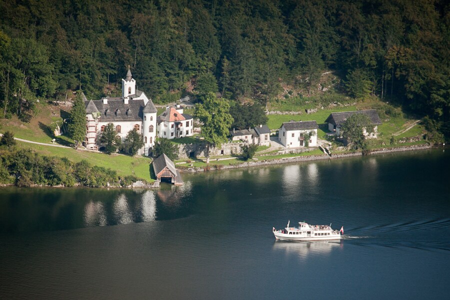 A serene lakeside view features a boat gliding on calm waters, with a charming historical building nestled among lush greenery on the shore. The scene reflects the peaceful surroundings, accentuated by the forested hills in the background.