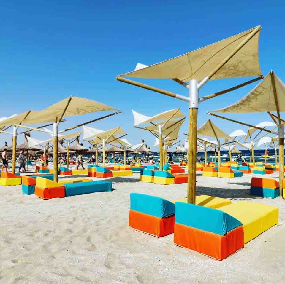 Colorful lounge chairs are arranged beneath large umbrellas on a sandy beach. The vibrant blue, yellow, and orange cushions provide comfort for sunbathers, while the clear blue sky enhances the inviting beach atmosphere.
