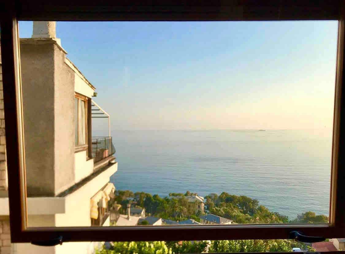 A wide window frames a panoramic view of the tranquil sea and coastal landscape. Gentle waves stretch into the horizon, reflecting soft sunlight. Below, lush greenery is visible, enhancing the serene atmosphere and connecting the interior space to the beautiful surroundings.