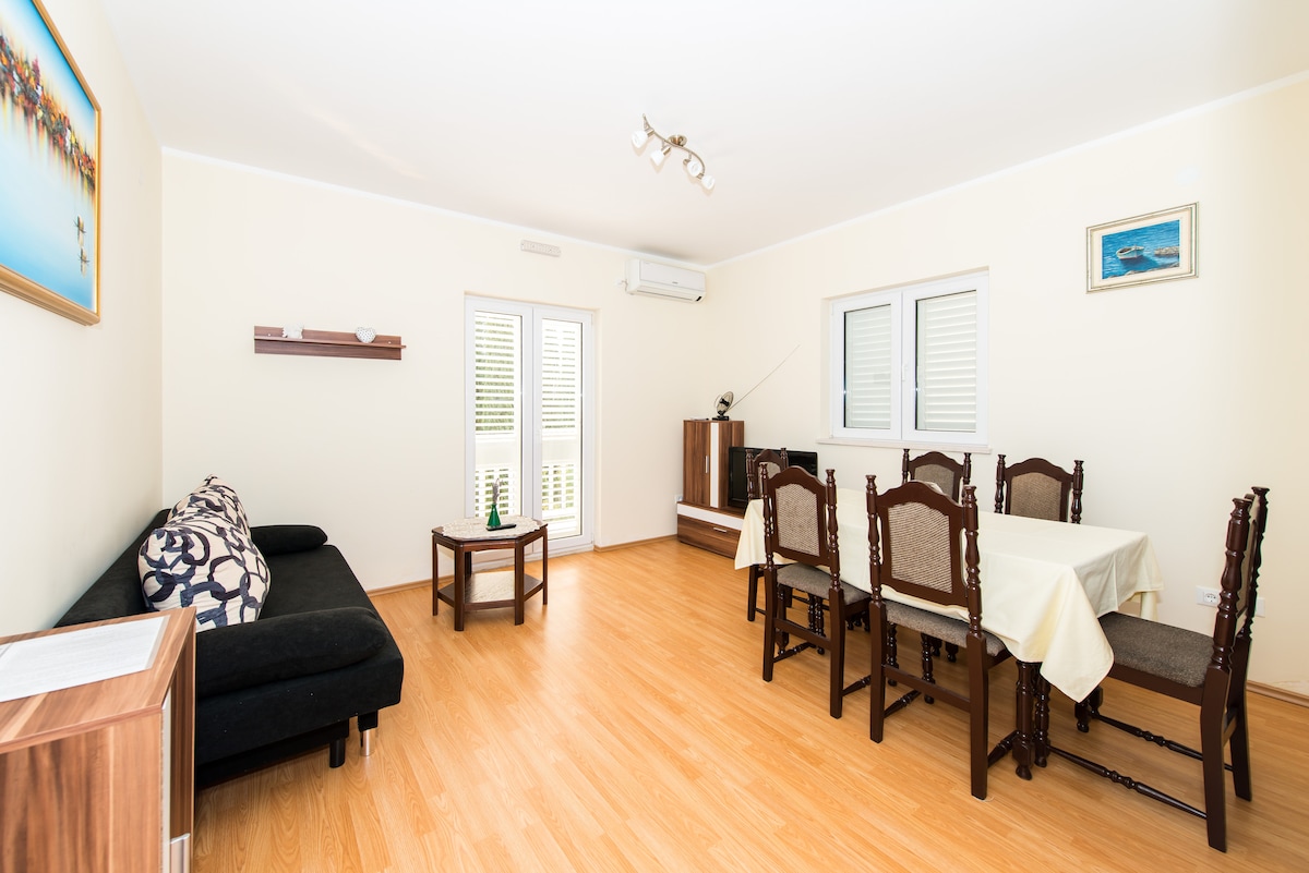 A light-filled living and dining area features a dark sofa and a wooden dining table surrounded by six matching chairs. Large windows provide views of the outside, while a piece of wall art adds a touch of decor. The space is complemented by wooden flooring.
