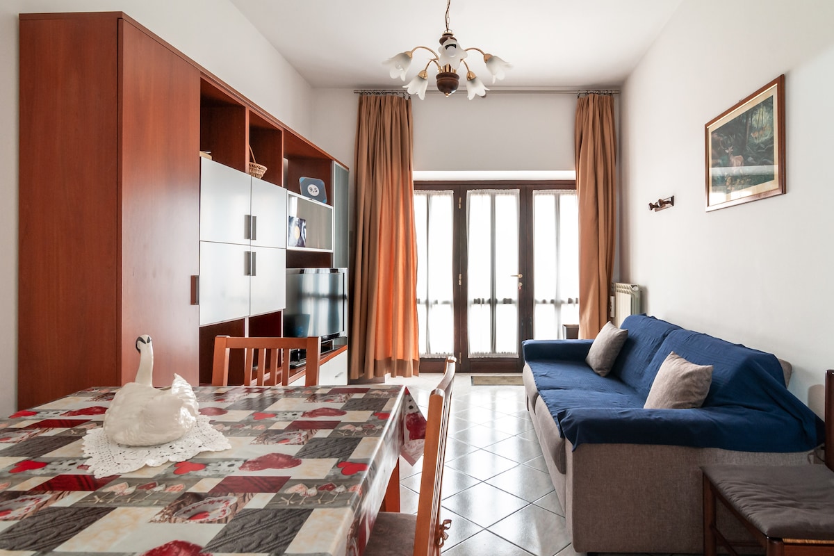 A living area features a dining table with a decorative tablecloth and a white swan figurine at its center. A blue sofa is positioned next to a wooden cabinet, complemented by large windows adorned with cream-colored curtains that allow natural light to fill the space.