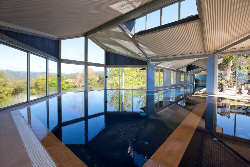 An indoor heated pool is surrounded by expansive glass walls that provide views of lush greenery outside. The water is calm and reflects the natural light streaming into the space, while the ceiling features a modern design with angled roofing.