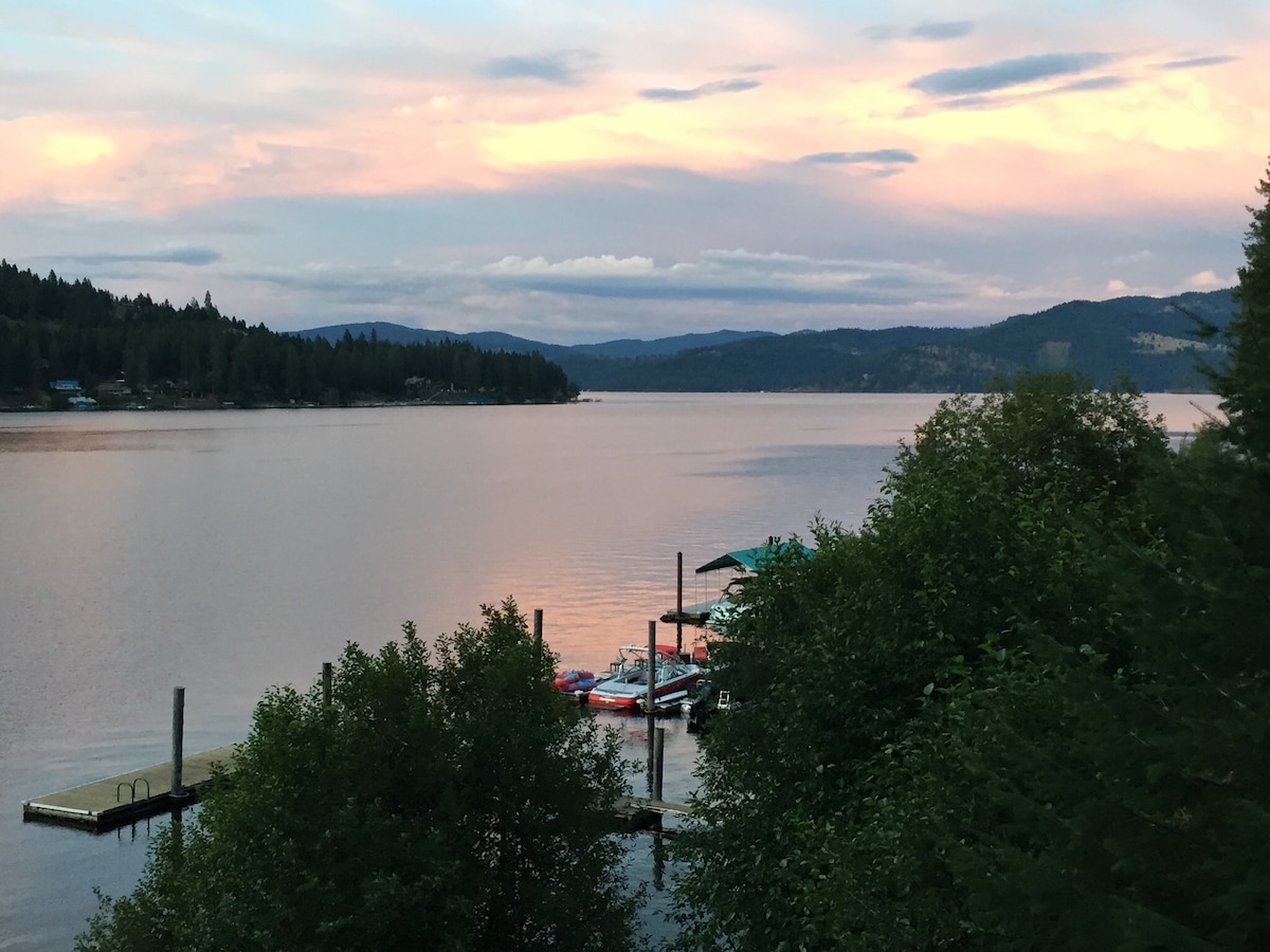 A serene view of Rockford Bay is captured at sunset, showcasing calm waters reflecting soft hues of pink and purple. A dock is visible, lined with boats, while the surrounding trees add a natural element to the picturesque lakeside setting.