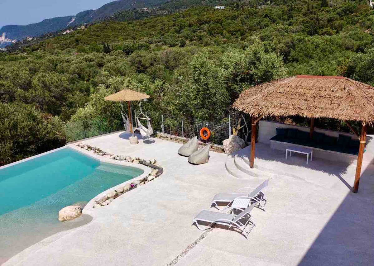 An inviting outdoor area features a luxury pool surrounded by a spacious sun deck. Two loungers are positioned beside the pool. A thatched-roof gazebo offers shaded seating, while a natural stone border accents the landscaped greenery in the background.
