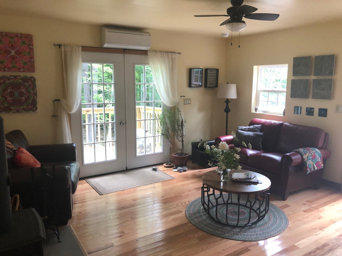 An open living area features hardwood floors and a ceiling fan. Multiple artworks adorn the walls, with two french doors allowing natural light to enter and providing access to an outdoor deck. A cozy seating arrangement includes a red sofa and a circular coffee table with floral decor.