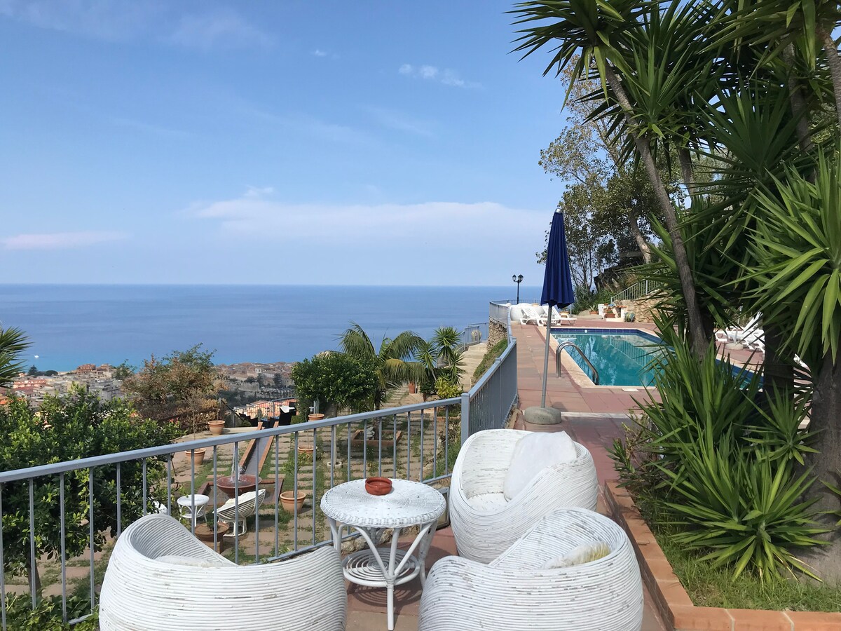 A terrace overlooking the sea is furnished with white lounge chairs and a small table. The vibrant blue pool can be seen nearby, surrounded by palm trees and Mediterranean greenery. The landscape includes distant views of the coastal town and expansive waters.