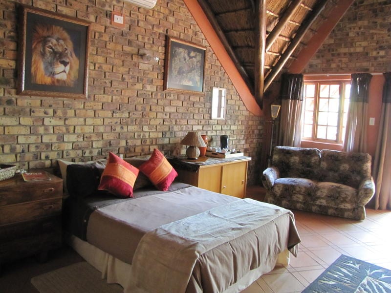 A spacious room features a large bed adorned with decorative cushions. Natural brick walls create a rustic backdrop, complemented by wooden furniture. An inviting seating area with a patterned couch is visible. Large windows allow light to enter, and artwork enhances the walls.
