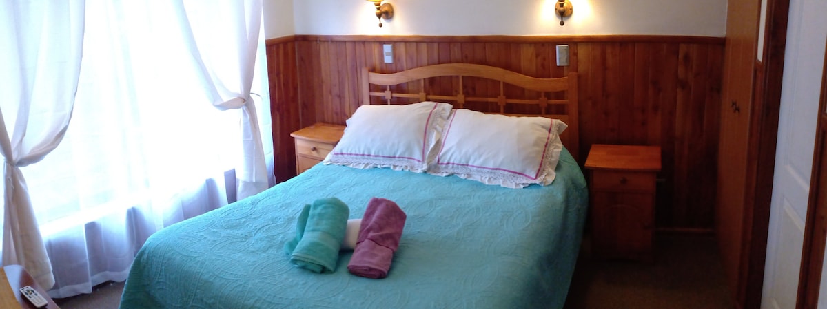 A bright sleeping area features a spacious bed with a light blue quilt and two pillows. Soft towels in mint green and lavender are neatly placed at the foot of the bed. Natural light enters through sheer curtains, softly illuminating the wooden accents on the walls.