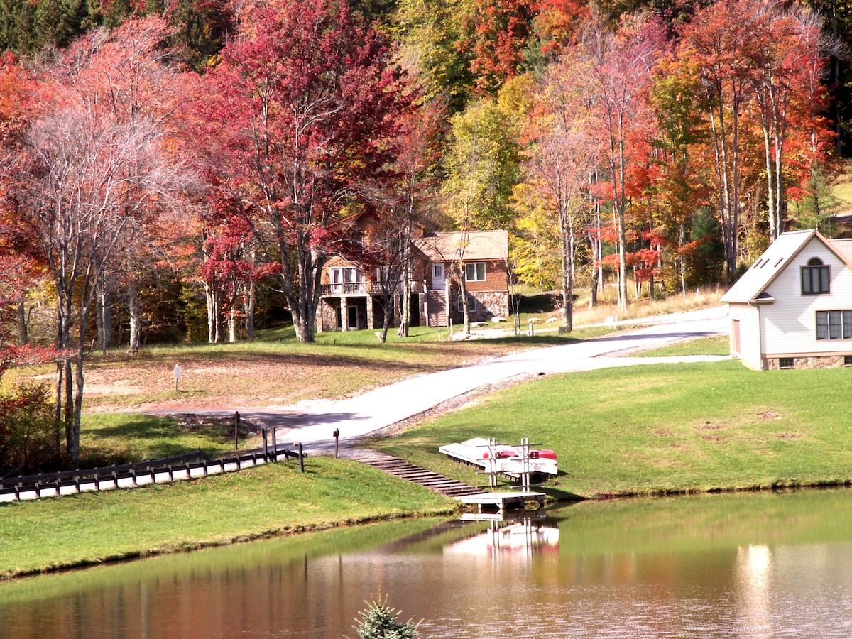 A serene view of a lake surrounded by lush greenery and vibrant autumn foliage. Two homes are situated near the water, with a dock featuring rowboats. The scene captures the peacefulness of nature in a colorful setting.