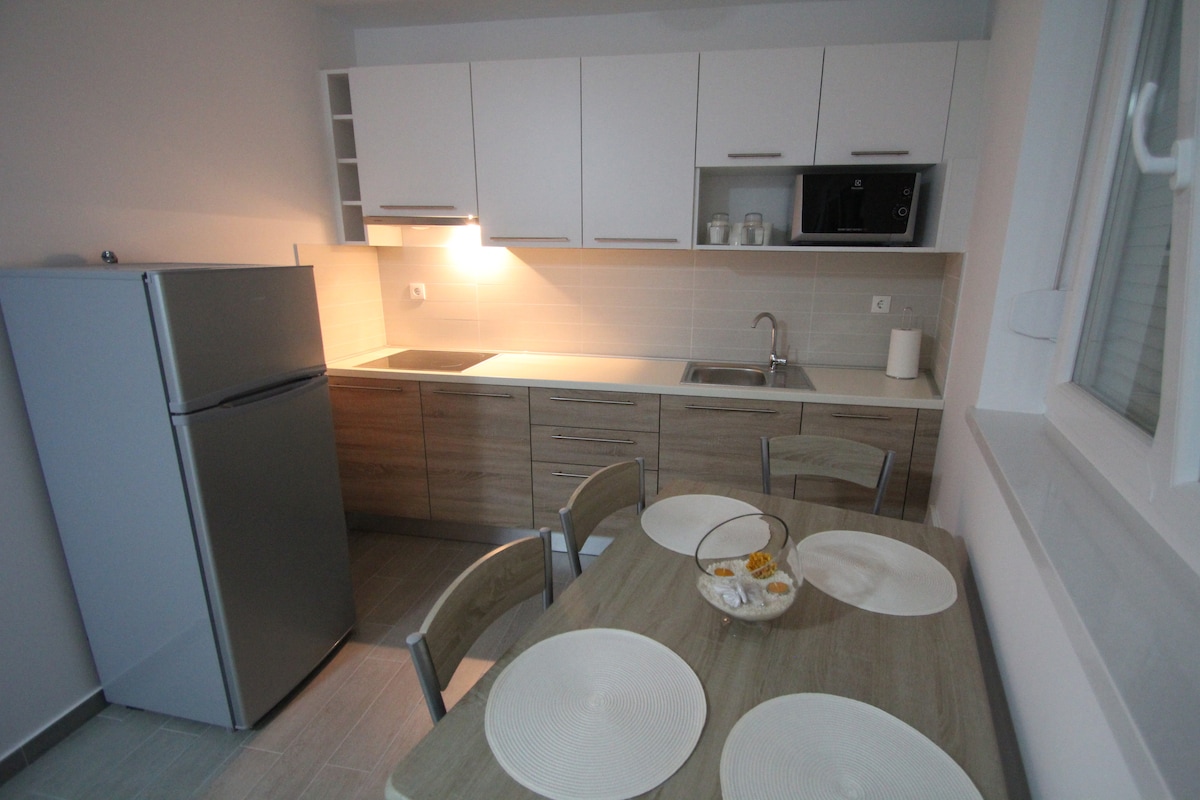 A modern kitchen area is presented, featuring sleek cabinetry and a stainless steel refrigerator. A wooden table is set with four place settings, complemented by a bowl of fruit. A microwave and sink are conveniently arranged, with soft lighting enhancing the functional space.