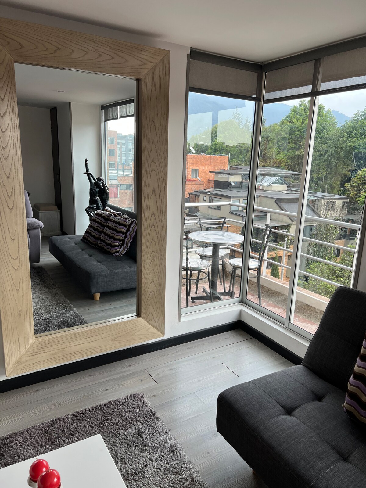 A modern living area is featured, showcasing a large mirror and a dark sofa adorned with decorative pillows. Large windows provide natural light and a view of the outdoors, with a small balcony furnished with a table and chairs, surrounded by greenery.
