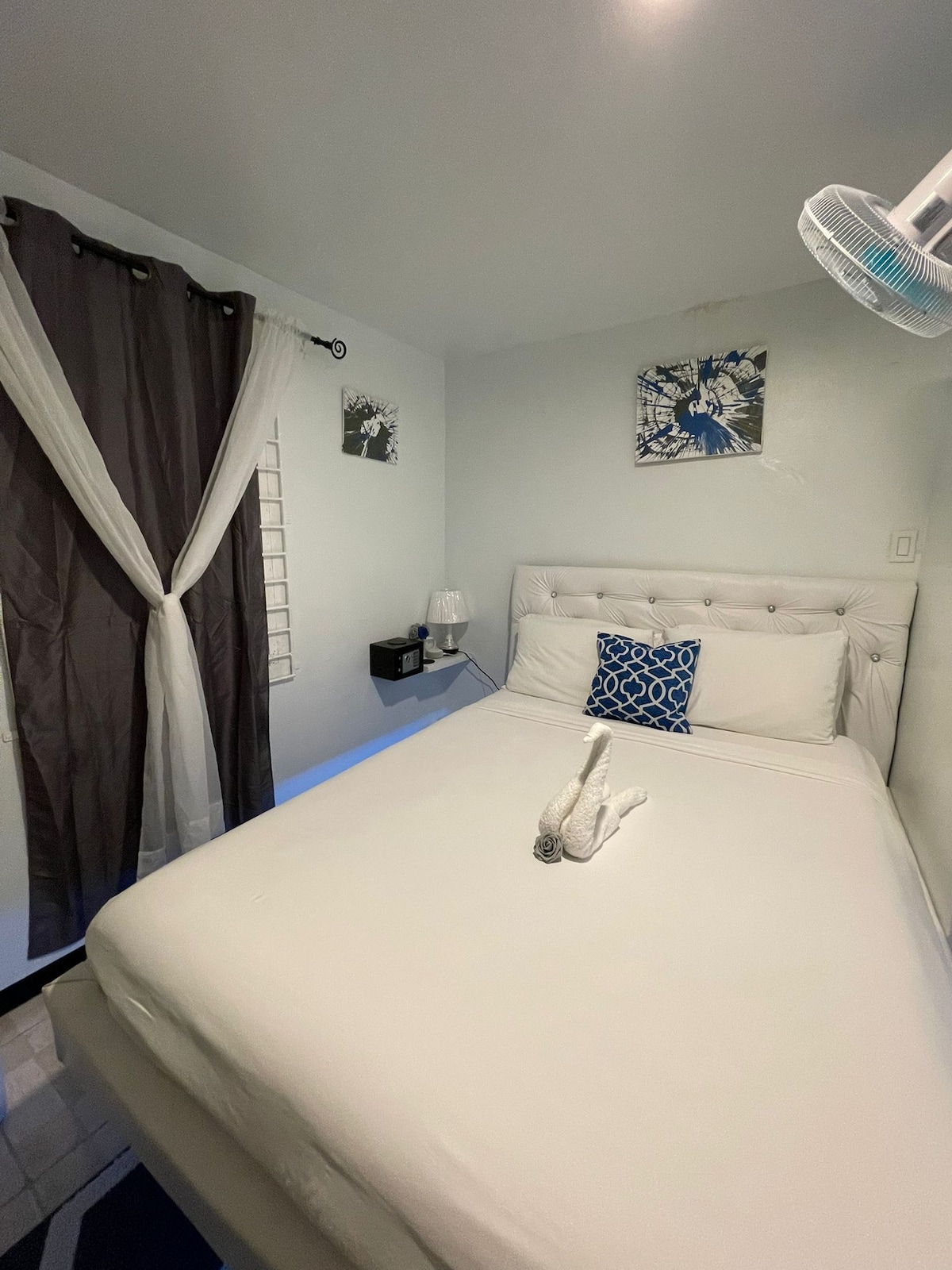 A compact bedroom features a spacious bed with light-colored linens and an accent pillow. A bedside lamp and a small nightstand are positioned next to the bed. Dark curtains frame the window, and two pieces of wall art add a touch of decor.