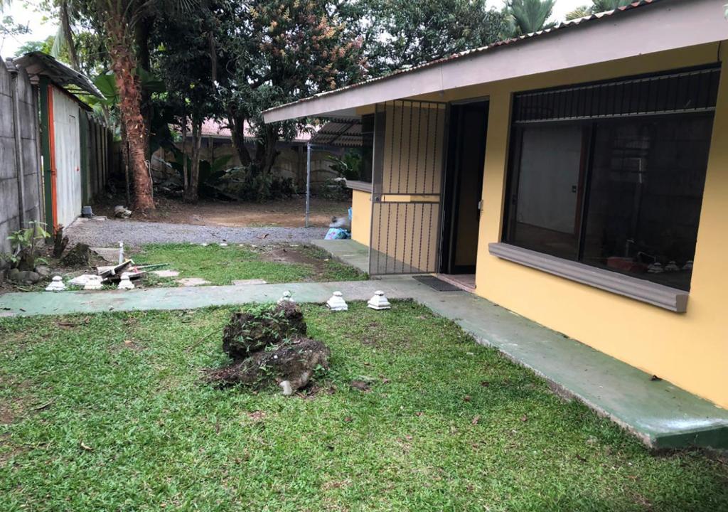 An outdoor view of the property reveals a well-maintained green lawn, complemented by a stone pathway leading to the entrance. A bright yellow wall contrasts with the surrounding nature, while large windows allow for natural light to enter the interior.