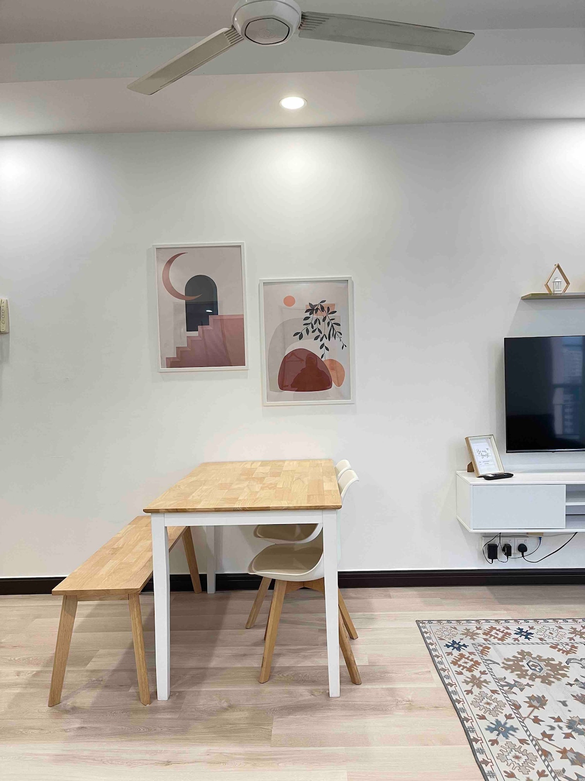 A dining area is presented with a light-colored wooden table and a matching bench, positioned against a neutral wall. Colorful artwork is displayed above the table, and a television is mounted on the adjacent wall, contributing to a modern and functional space.