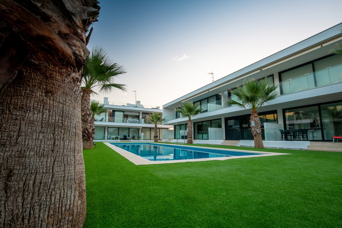 A modern apartment complex is framed by lush palm trees and manicured grass, featuring a rectangular swimming pool at its center. Large glass windows line the building, offering views of the pool area. The tranquil setting is enhanced by the evening light.