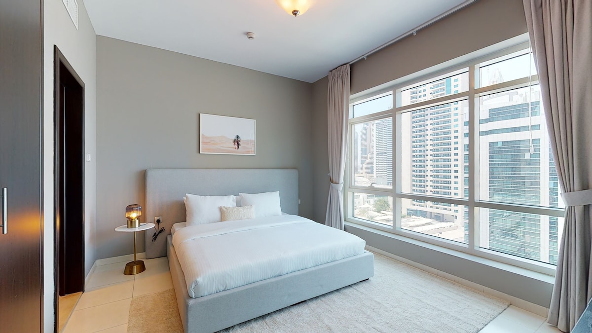 A minimalistic bedroom features a spacious bed against a neutral wall, with soft linens and pillows providing a restful setup. Large windows dominate one side, offering views of the cityscape. Soft curtains frame the windows, allowing natural light to brighten the room.