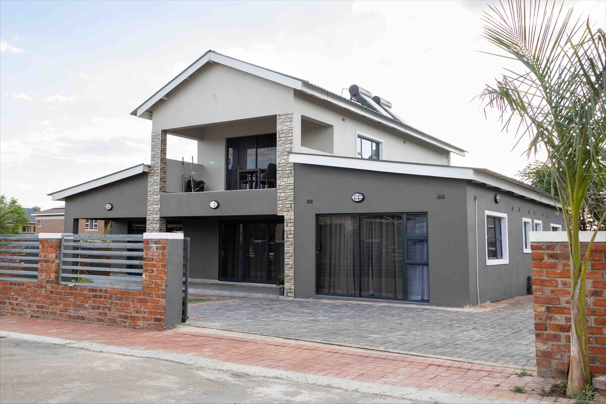 The modern two-story villa is presented with a contemporary design and features a stone-accented facade. Large glass doors offer views of the outdoor patio space, while a secure brick wall provides privacy. A lush palm tree is visible, enhancing the neighborhood's greenery.