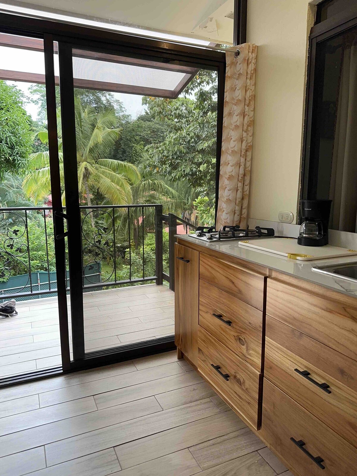 A modern kitchen area features light wooden cabinets and a countertop. A large glass door opens to a balcony surrounded by lush greenery. A small coffee maker is positioned on the counter, and a gas stove is present alongside the sink.