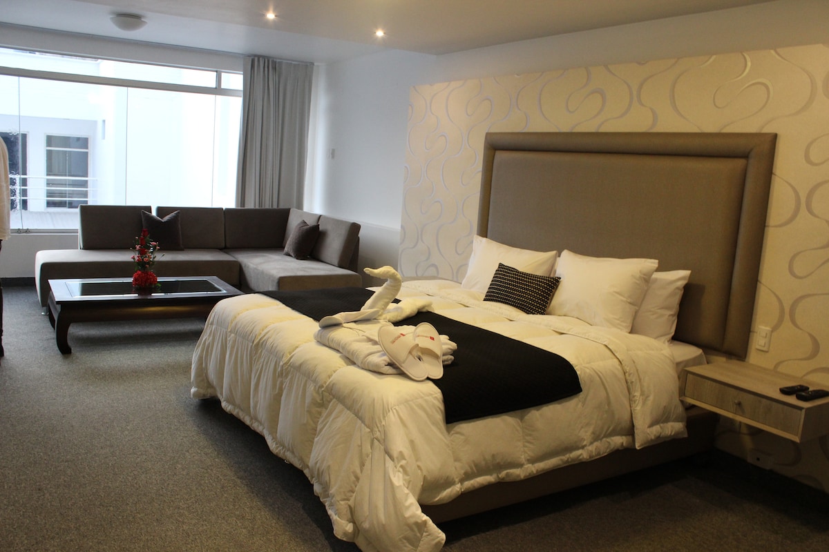 A spacious room is presented with a large bed featuring quality bedding. A modern sofa and coffee table are arranged in a separate seating area, while natural light filters through a large window. Decorative elements include a small vase with flowers on the bedside table.