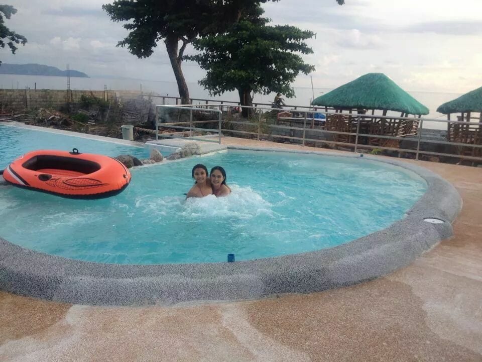 A swimming pool is nestled within a resort setting, surrounded by lush greenery and beach views. A bright orange float is visible in the crystal-clear water. Thatched-roof huts are positioned nearby, providing shaded seating areas, inviting relaxation by the water during a cloudy day.
