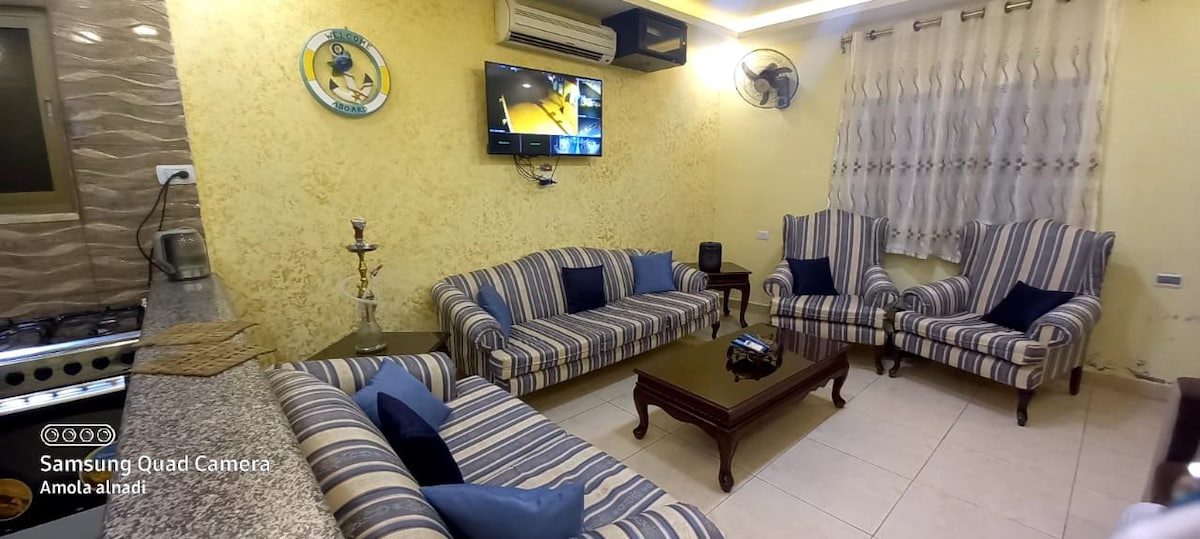 A living area features striped sofas positioned around a wooden coffee table. A flat-screen television is mounted on the wall, complemented by a decorative wall piece. Natural light filters in through the window, which is dressed with sheer curtains.