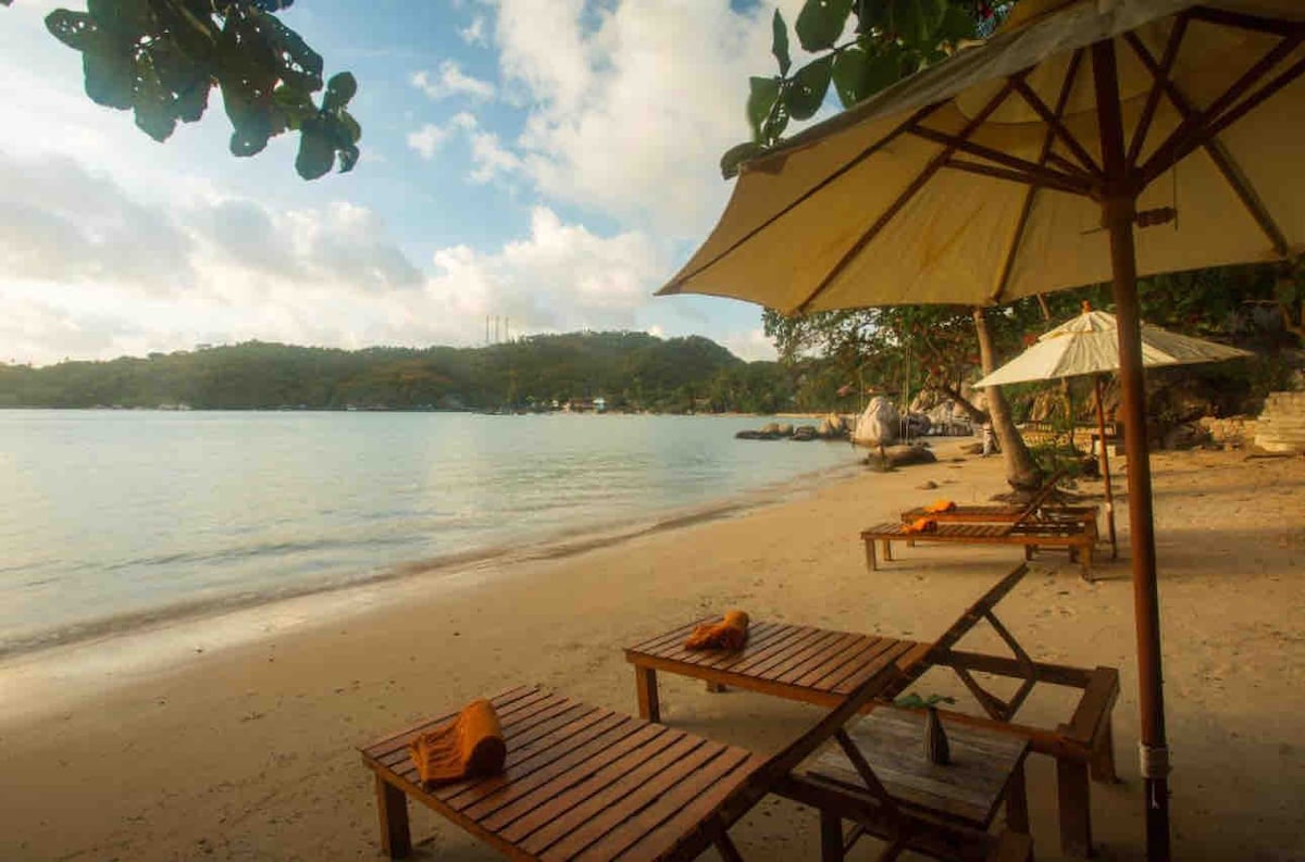 Two wooden lounge chairs are positioned on a sandy beach beneath a large umbrella, offering a serene view of the calm water and distant hills. Soft clouds are visible in the sky, and the shoreline is framed by lush greenery.