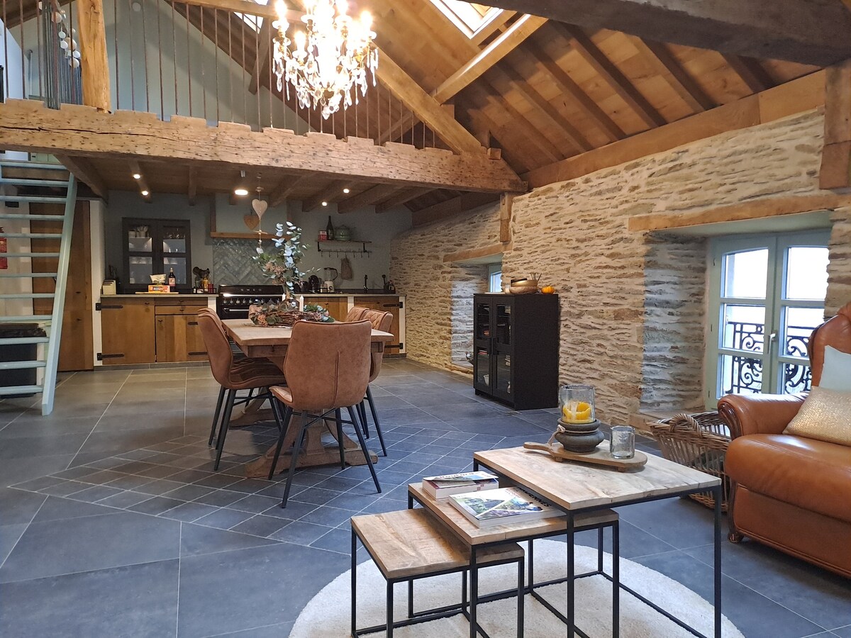 A spacious living area features high ceilings with wooden beams, enhanced by natural light from large windows. A stylish dining table with leather chairs sits at the center, accompanied by a cozy seating arrangement. An elegant chandelier adds charm to the rustic ambiance.