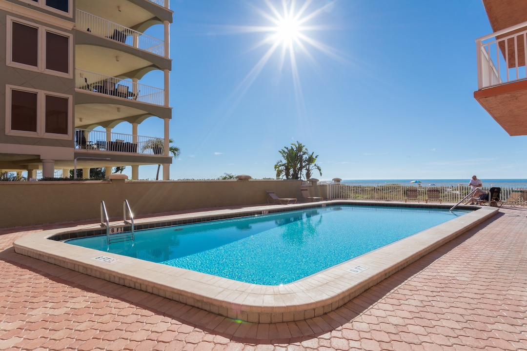 A clear blue sky is visible above a sparkling pool, with sunlight glinting off the surface of the water. The pool area is framed by beige stone tile and bordered by lush greenery, while the ocean is seen in the background, enhancing the tranquil atmosphere.