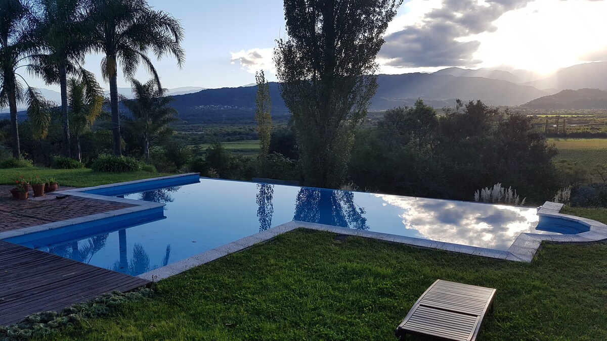 An infinity pool reflects the sky and surrounding landscape, offering a view of distant mountains and open fields. Lush green grass borders the pool area, complemented by wooden decking and a lounge chair positioned nearby under the gentle glow of evening light.
