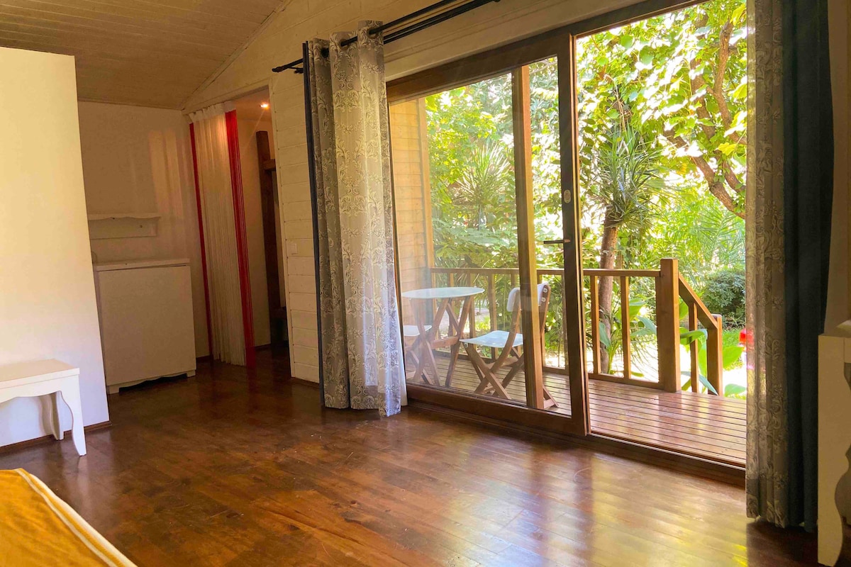 A spacious room features large glass doors that open to a private deck surrounded by lush greenery. Wooden flooring complements the light-colored walls. Natural light filters through sheer curtains, creating a warm atmosphere. A small table and chairs are visible on the deck.
