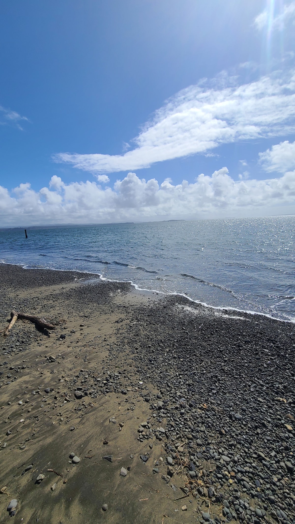 A serene shoreline is depicted, showcasing a sandy beach with scattered pebbles. Gentle waves lap against the shore, and the tranquil waters reflect the blue sky and fluffy white clouds. A distant post can be seen rising from the water.