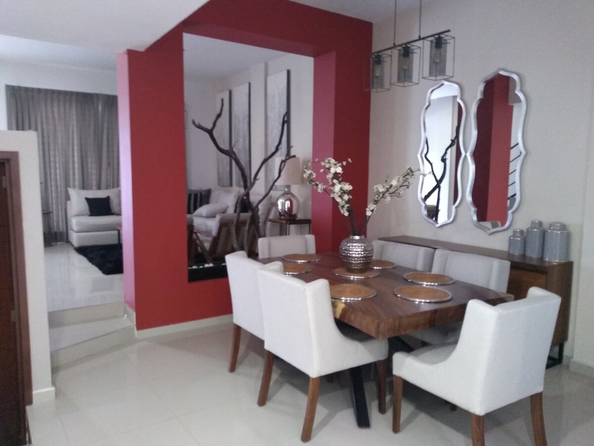 A dining area features a wooden table surrounded by six upholstered chairs. Decorative items, including a vase with flowers, add elegance. The space transitions into a spacious living area, visible beyond a mirror, where soft furnishings create a comfortable setting.