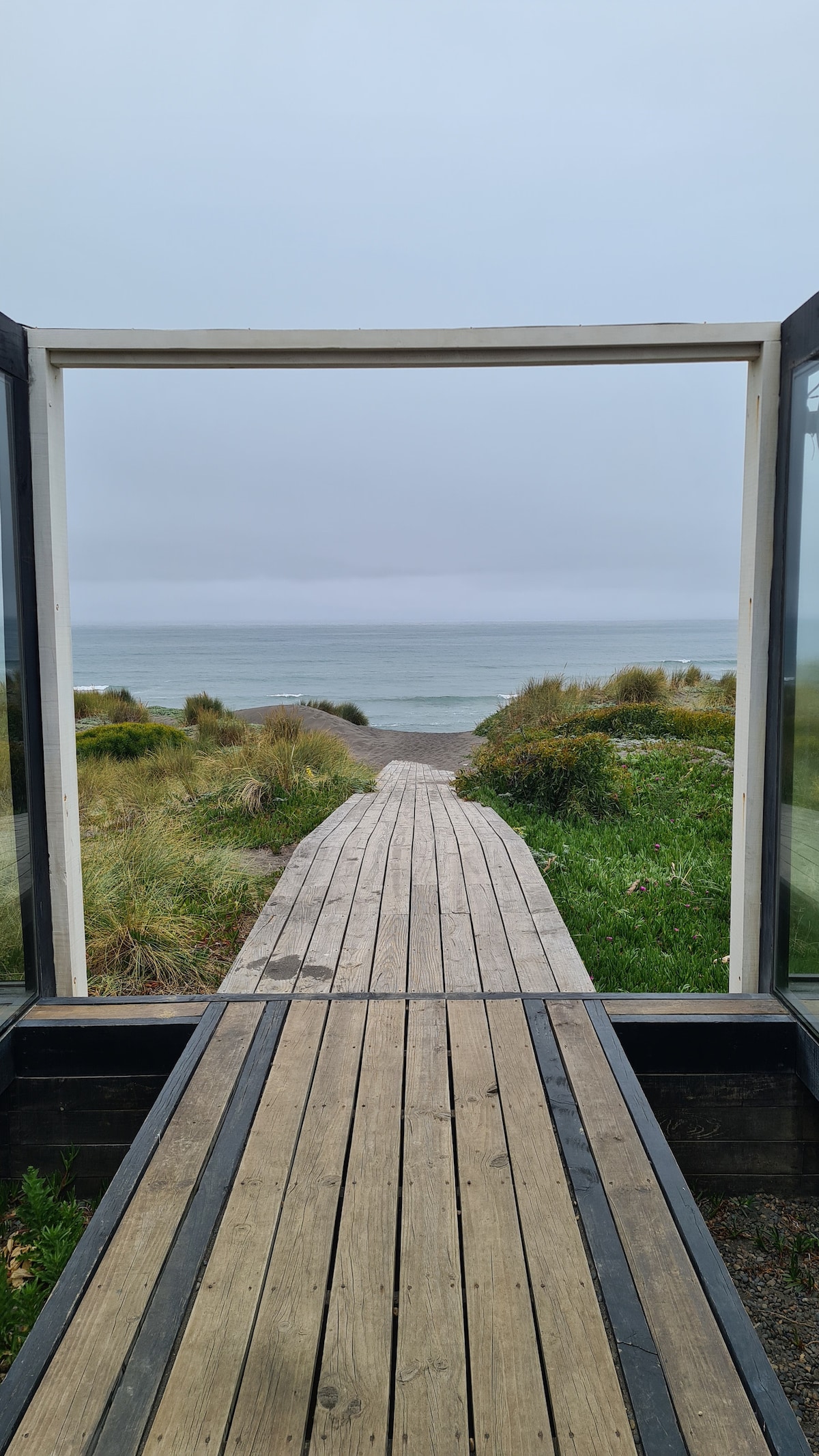 A wooden pathway leads through a glass entryway, directing attention towards the ocean. Green grass and scattered shrubs frame the path, which stretches towards the tranquil water and cloudy sky in the distance.