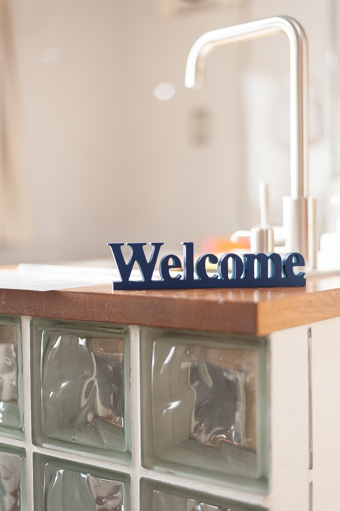 A kitchen counter is displayed with a decorative 'Welcome' sign positioned prominently. The counter features a wooden top complemented by a mosaic of glass block accents, while a sleek faucet is visible in the background, enhancing the inviting atmosphere of the space.