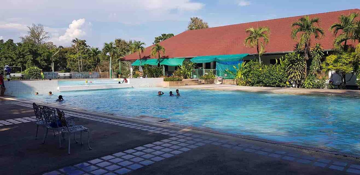 A resort-style swimming pool is surrounded by lush greenery and tropical plants. People are seen enjoying the water, while shaded seating areas are provided nearby. The building in the background features a red roof and expansive outdoor space, creating a relaxed atmosphere.