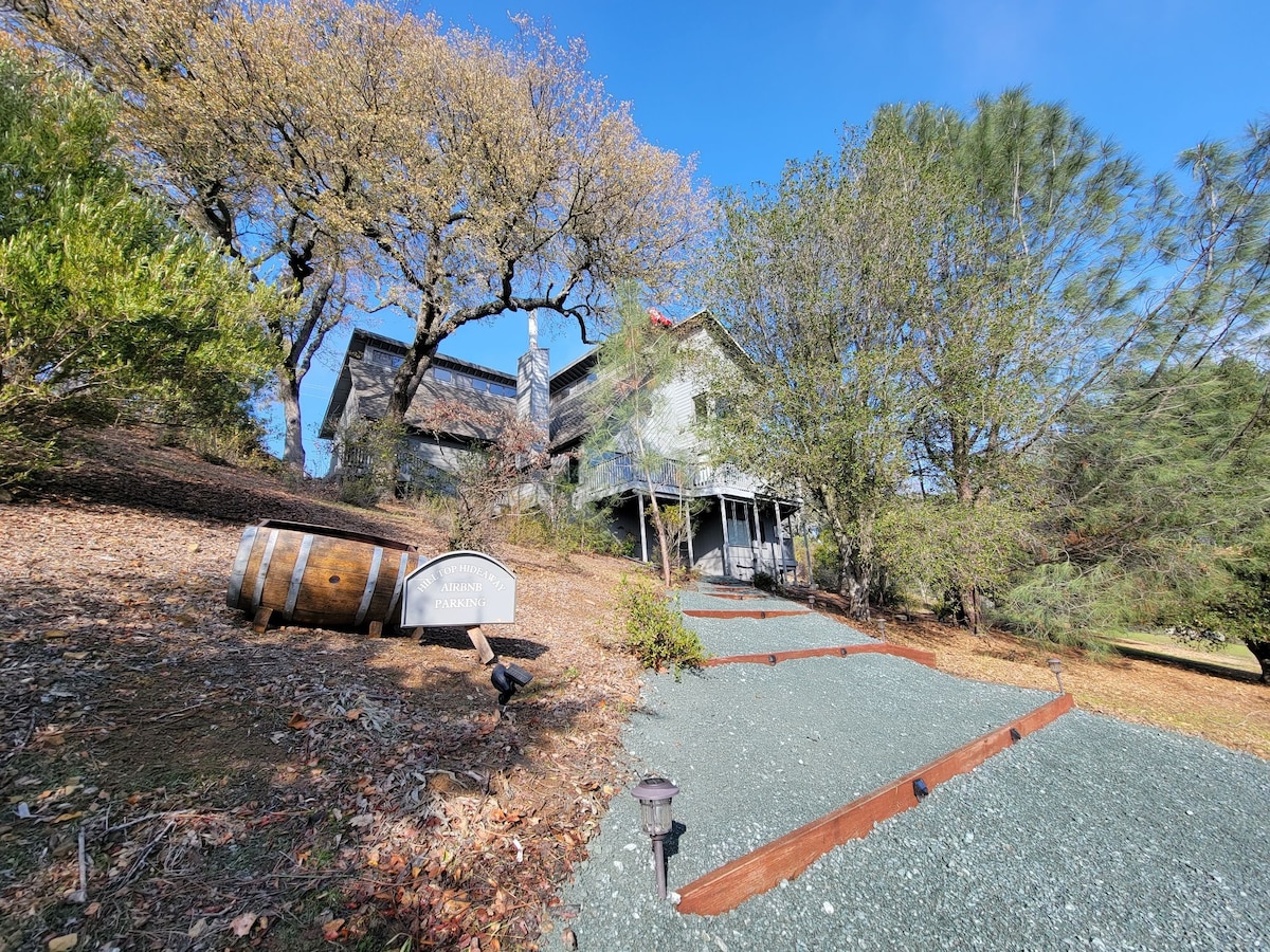 A scenic view of the guest suite's exterior is presented, featuring a gently sloped pathway leading to the entrance. Surrounding greenery and mature trees enhance the natural setting, while a decorative wooden barrel adds a rustic touch to the landscape. Clear blue skies are observed above.
