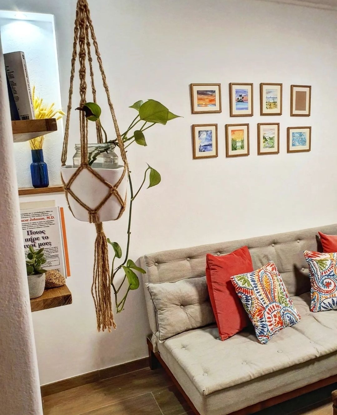 A cozy living area displays a comfortable sofa adorned with colorful cushions. A wall-mounted shelf holds decorative items and plants, while framed artwork is arranged neatly above. Soft lighting enhances the inviting atmosphere of the space, which is visually clean and well-organized.