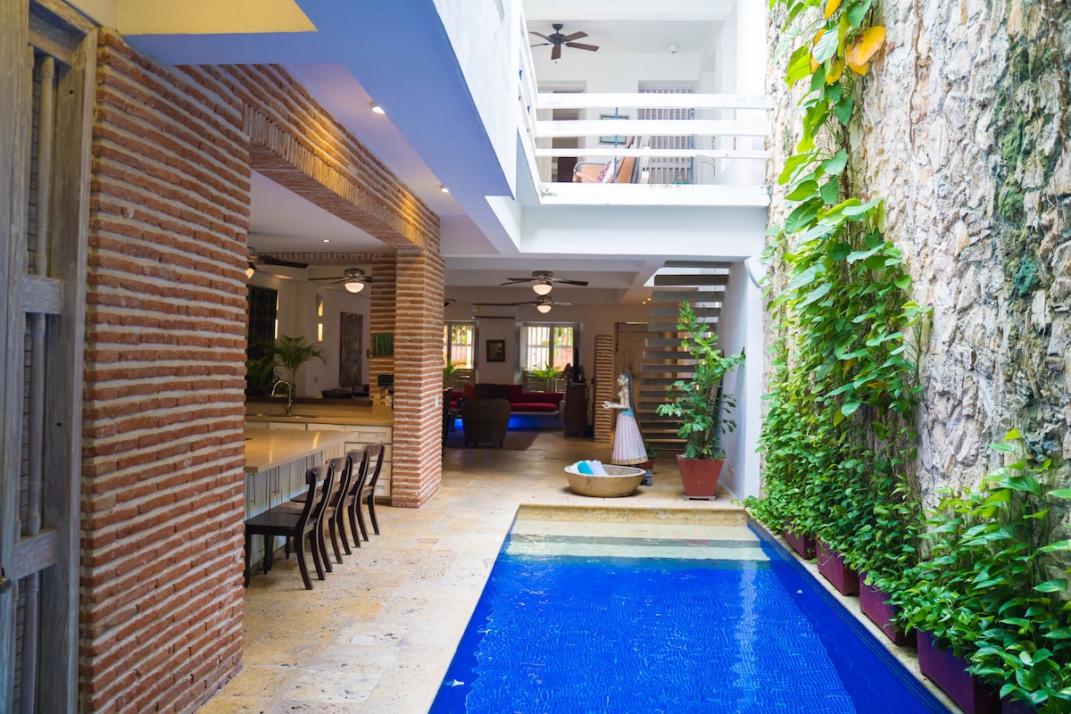 An indoor pool is positioned between brick walls, surrounded by lush greenery. Natural light floods the area, highlighting the living space, which includes a dining area with multiple chairs and an open staircase leading to an upper level. The tranquil water reflects the surrounding details.
