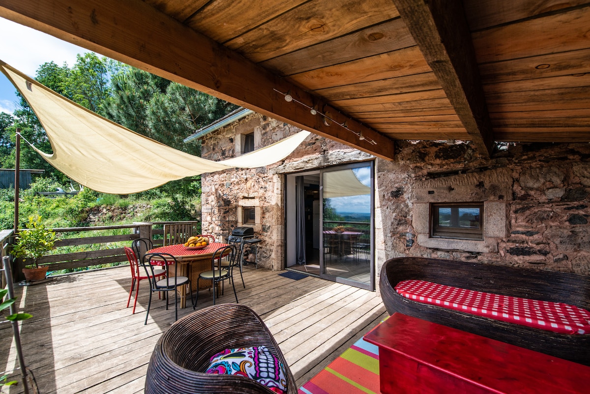 A wooden deck is adorned with a seating area featuring cozy lounge chairs and a round dining table. Colorful textiles and fresh fruit on the table add vibrancy. A spacious entrance leads to the interior through sliding glass doors, with natural surroundings visible in the background.