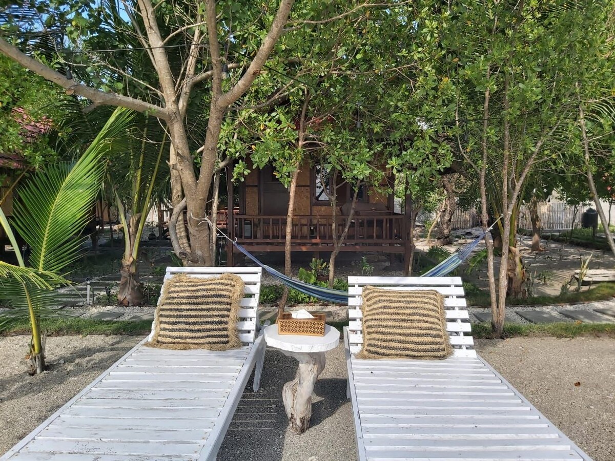 Two white loungers are positioned on a sandy area, with decorative cushions resting atop them. A small round table stands between the loungers, complemented by a basket. In the background, a bungalow with wooden details is surrounded by lush greenery and a hammock is seen hanging among the trees.