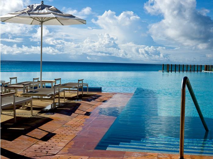 An inviting infinity pool meets the clear blue waters of the Caribbean Sea. Comfortable lounge chairs are arranged under a large umbrella, offering shade. The sky is adorned with scattered clouds, reflecting the tranquil atmosphere of the resort.