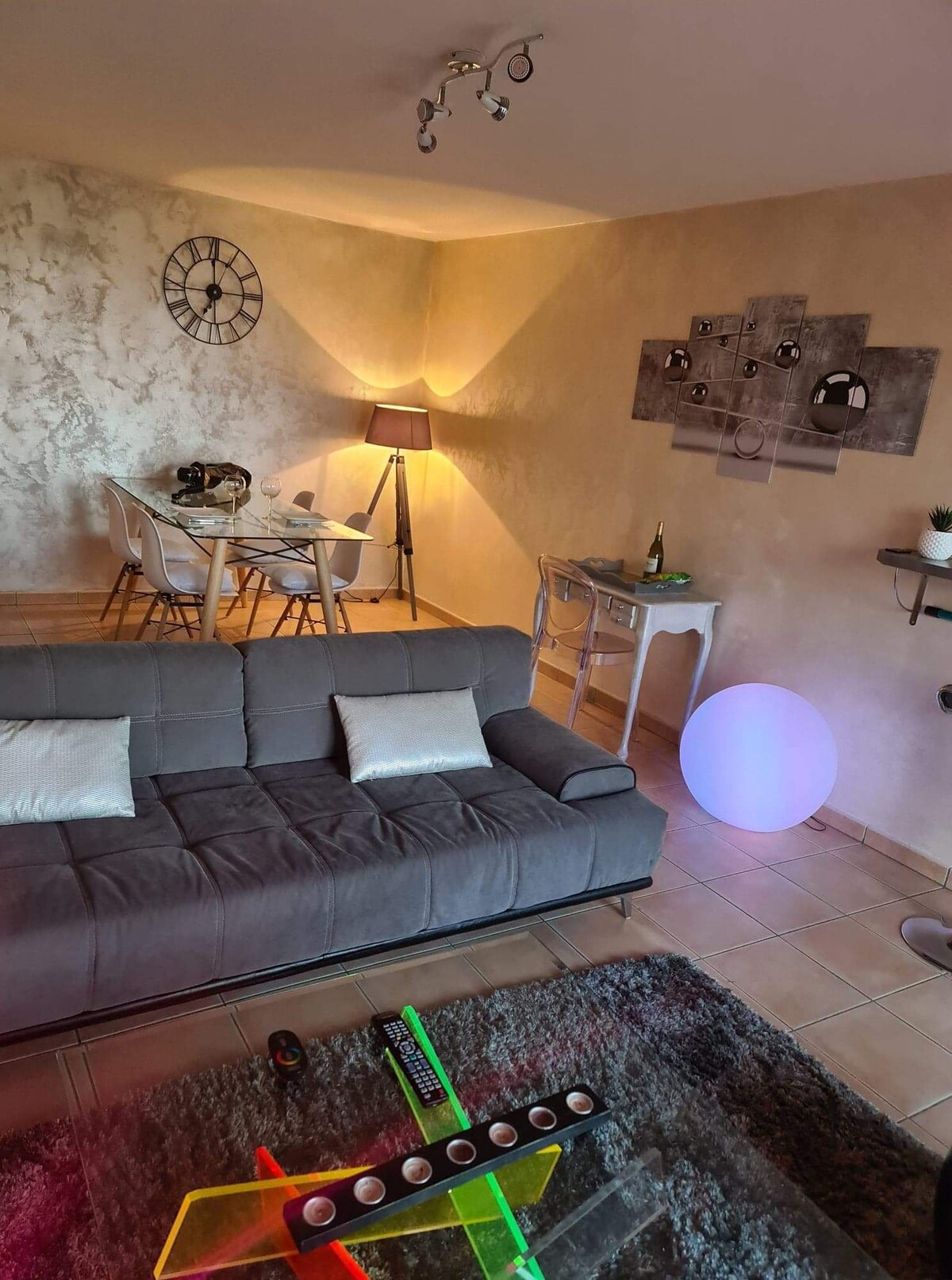 The living area features a gray sofa paired with decorative pillows. A dining table is set for four, complemented by a stylish lamp. A colorful light source adds to the ambiance, while wall art and a clock enhance the room's decor.