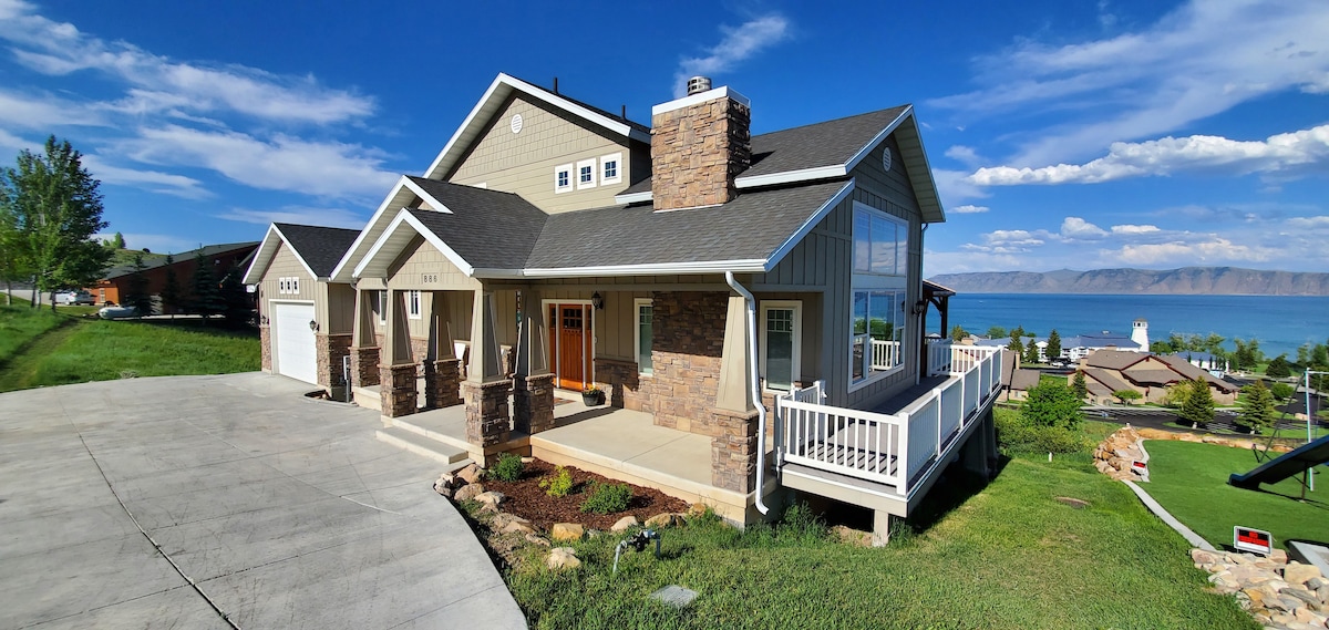 A craftsman-style home is featured, set against a clear blue sky with scenic views of Bear Lake in the background. A spacious front porch is visible, accompanied by well-maintained landscaping and a driveway leading up to the entrance.