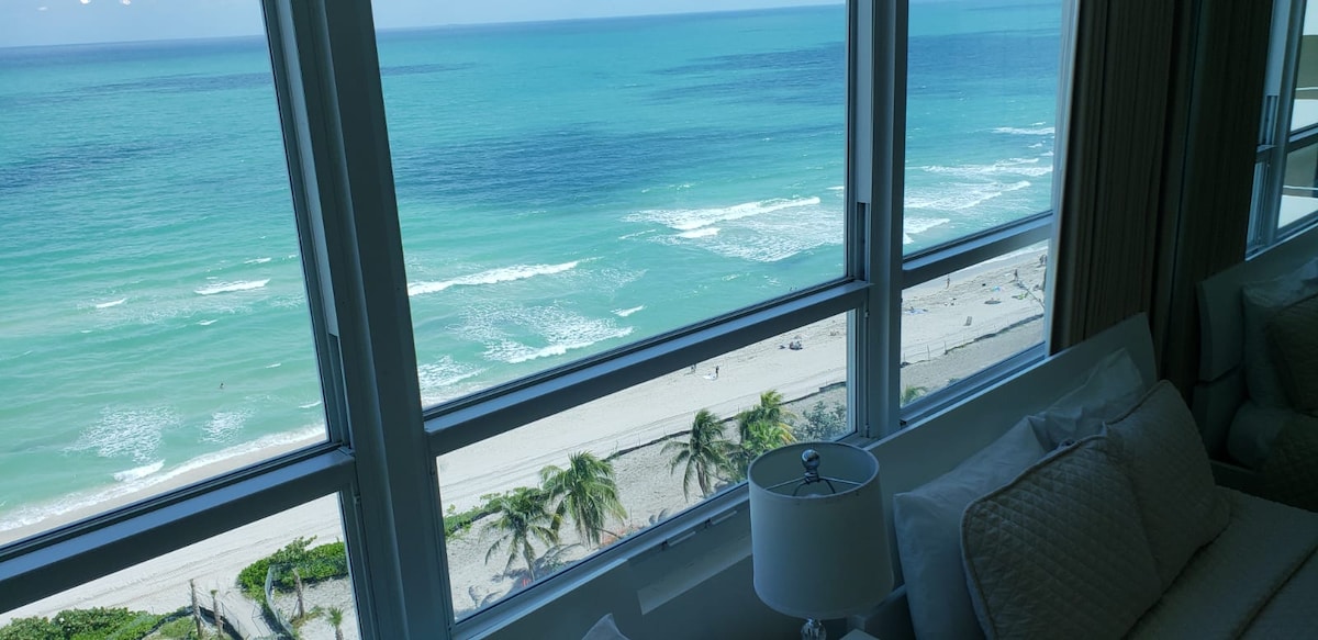 A view of the ocean is framed by large windows, showcasing vibrant blue waters and gentle waves. The coastline is visible below, with sandy beaches and palm trees lining the shore. Natural light brightens the space, creating a serene atmosphere.