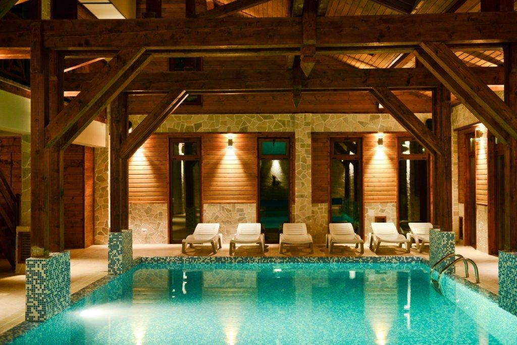 An indoor pool is framed by wooden beams and natural stone walls. Lounge chairs are arranged around the water, illuminated by soft lighting from the surrounding windows. The turquoise pool surface reflects the warm tones of the rustic architecture.