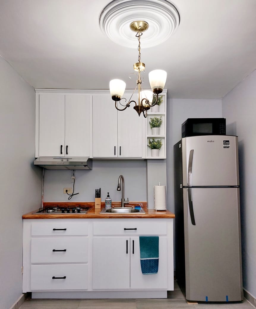 A compact kitchen area is designed with white cabinetry and wood countertops. A stove and kitchen sink are positioned next to a refrigerator with a metallic finish. A chandelier with three bulbs adds lighting, while small plants are displayed on an upper shelf.