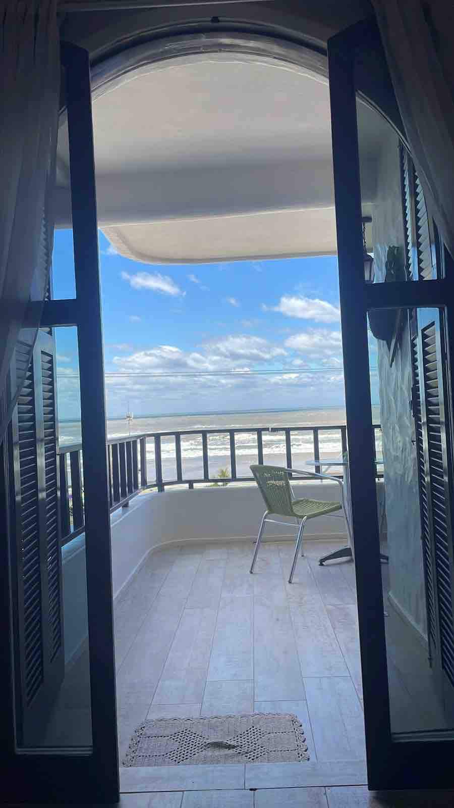 Double doors open to an expansive balcony overlooking the beach, with a single green chair positioned for ocean views. The bright sky is filled with scattered clouds, enhancing the sense of spaciousness and natural light.