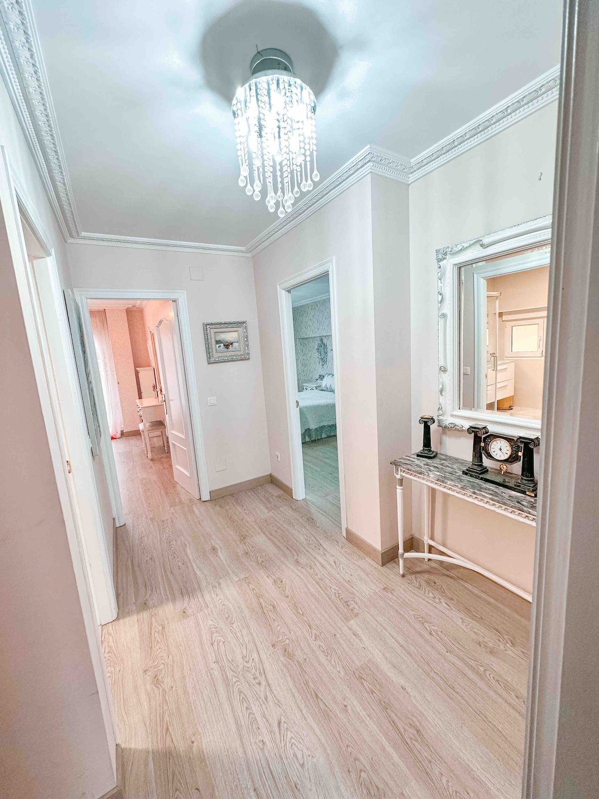 A well-lit hallway is seen, featuring a stylish chandelier overhead. A decorative mirror is mounted on the wall, reflecting the space. A small console table holds a clock, while light wood flooring extends down the corridor, leading to a doorway on the right.