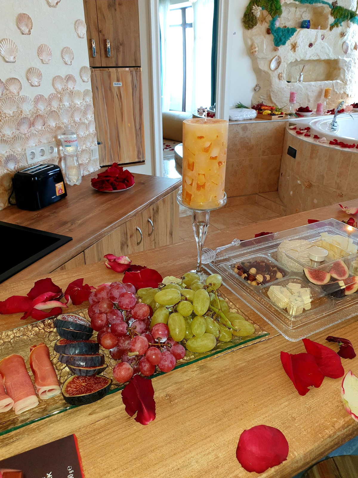 A wooden countertop displays an assortment of fresh grapes, figs, and cured meats, arranged with care. A glass container holds various snacks, accompanied by small bowls of sauces. Rose petals scatter across the surface, and a candle adds a warm glow to the scene.