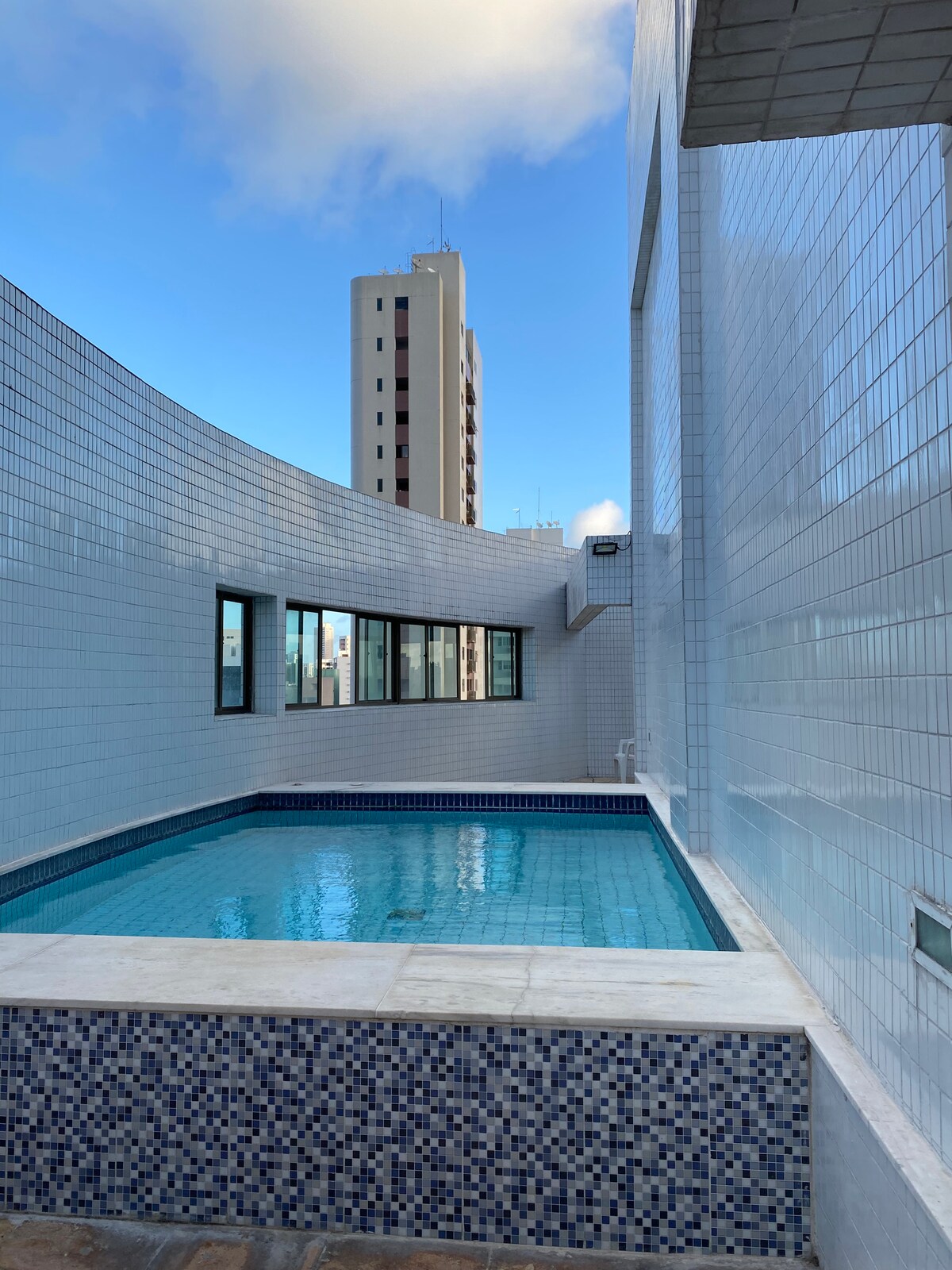 A rooftop pool is visible with clear blue water, surrounded by curved white tiled walls. Large windows provide a view of the cityscape, contrasting against the bright sky. The area is well-lit, promoting a serene atmosphere for relaxation.